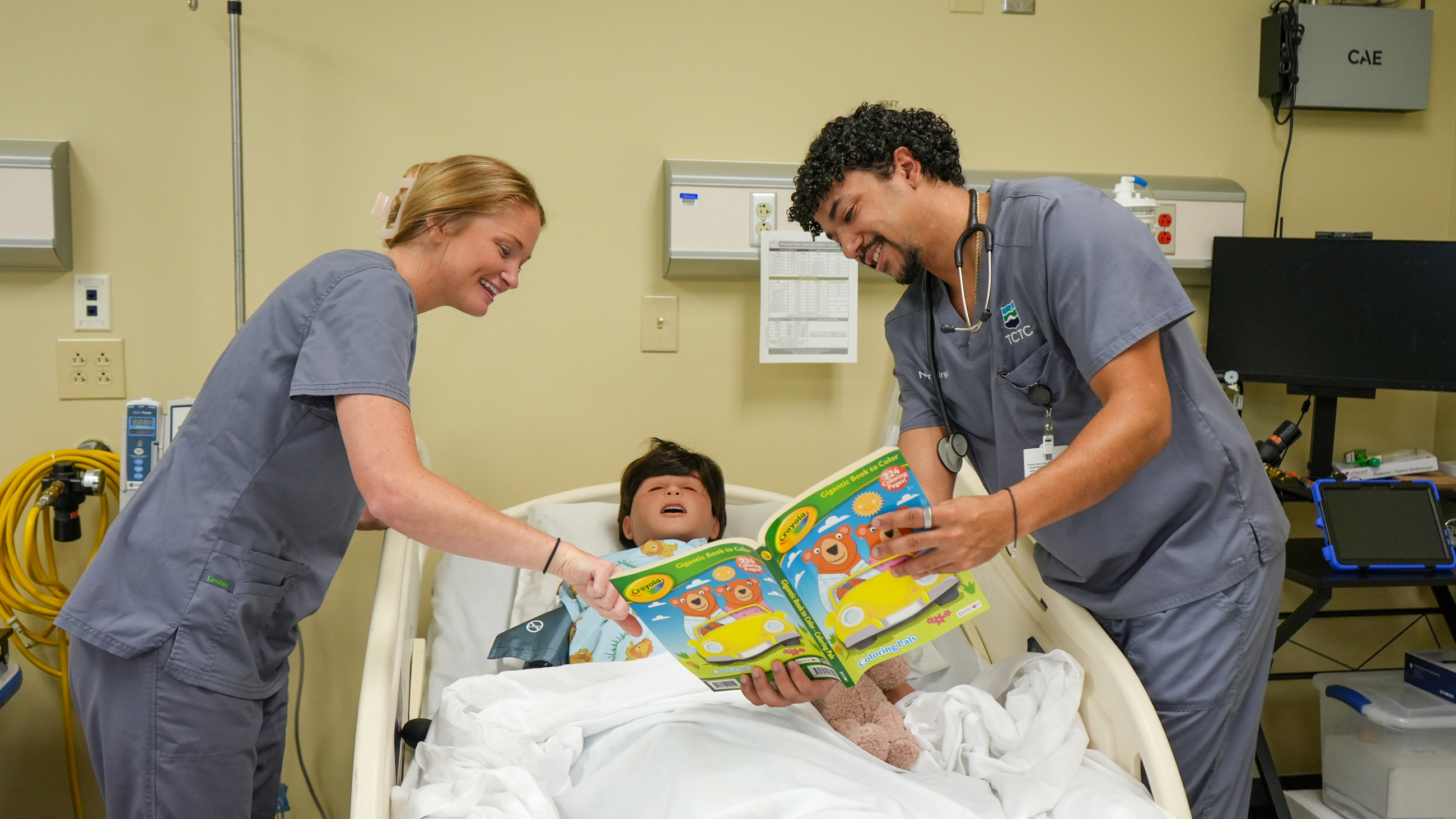 Two TCTC nursing student with a fake patient in a hospital bed reading them a book. 