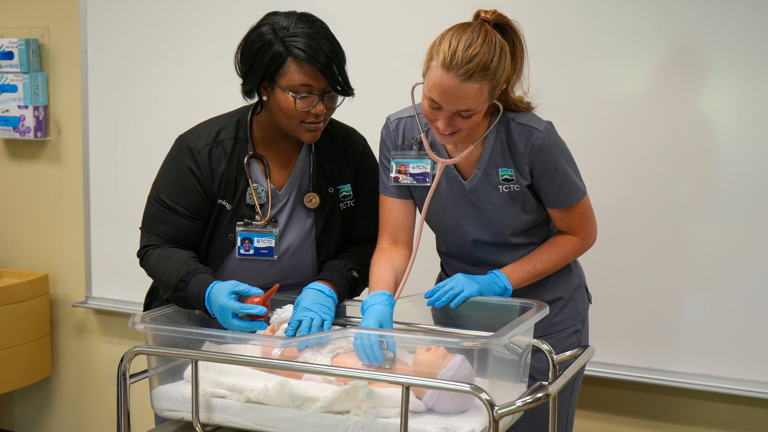 Two TCTC nursing students working over a fake baby in a bed checking their vitals. 